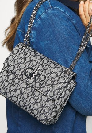 Borsa a tracolla con motivo bianco e nero e catena, con chiusura a pattina e accento del logo, appoggiata su uno sfondo in denim blu.