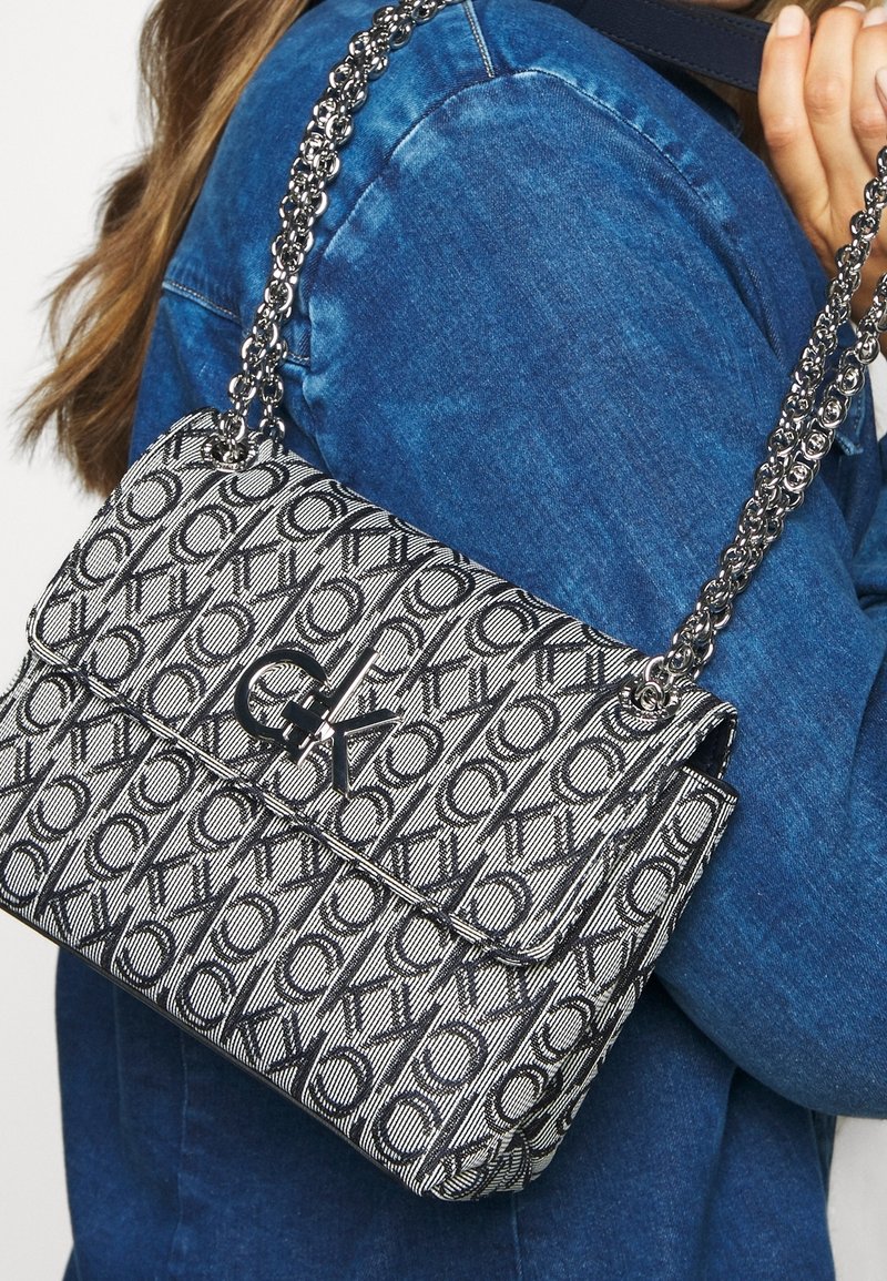 Borsa a tracolla con motivo bianco e nero e catena, con chiusura a pattina e accento del logo, appoggiata su uno sfondo in denim blu.