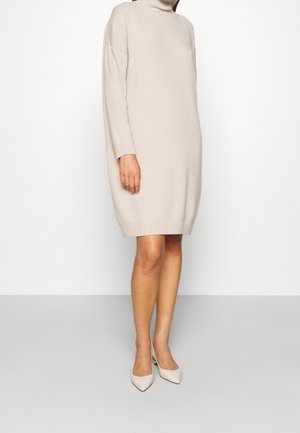 Kobieta ubrana w beżową sukienkę swetrową z długim rękawem i golfem sięgającą kolan oraz beżowe szpilki w szpic, stojąca na jednolitym tle.