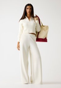 Maglione bianco a trecce con collo a zip, abbinato a pantaloni bianchi a gamba ampia. La modella tiene una borsa tote beige con dettagli bordeaux.