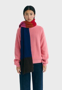 Pull en tricot rose à coupe lâche, col rond ; associé à une écharpe multicolore rayée avec des sections rouges, bleues et brunes.