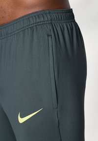 Tamnozeleni sportski hlače s teksturiranim završetkom, s žutim Nike logom i elastičnom strukom. Uključuje bočne džepove za funkcionalnost.