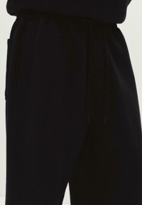 Detailní záběr černých tepláků s bočními kapsami a elastickým pasem se stahovacími šňůrkami.