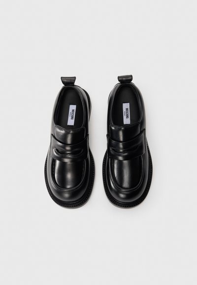 Melnu ādas loafers ar noapaļotiem purngaliem, dekoratīvu siksnu un markētiem zoles tagiem, izgatavoti uz balta fona.