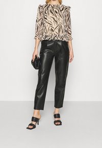 Beige Bluse mit schwarzem Tiermuster, Rüschensäumen und einem ausgestellten Saum, kombiniert mit schwarzen Kunstlederhosen und schwarzen Absätzen.
