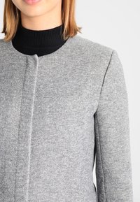Manteau gris en mélange de laine avec un col rond, des épaules structurées et un design minimaliste, présentant une texture subtile et sans fermetures visibles.