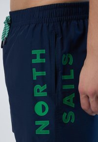 Shorts da nuoto navy con il testo verde "NORTH SAILS" e un logo testurizzato. Include un cordoncino verde e una tasca laterale. Materiale leggero.