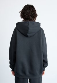 Personne avec des cheveux noirs mi-longs portant un sweat à capuche noir ample et uni, de dos contre un fond blanc.