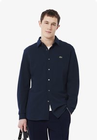 Marineblå langermet skjorte med knapper og spiss krage, med en liten grønn logo, glatt tekstur og subtile knappedetaljer.