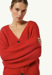 Rote gestrickte Strickjacke mit V-Ausschnitt, lockeren Ärmeln und grünen Knöpfen, die einen strukturierten Stoff und ein grob gestricktes Design aufweist.