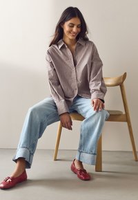 Jeune femme souriante, assise sur une chaise en bois, portant une chemise rayée taupe, un jean large bleu clair et des ballerines rouges.