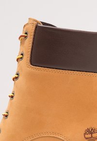 Bottine en cuir marron avec extérieur en daim beige, œillets ronds en métal, lacets jaunes et rouges, et détail de logo de la marque.