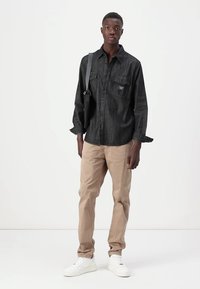 Black denim shirt with buttoned pockets, beige pants, and white sneakers. The shirt has a relaxed fit and long sleeves.