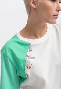 Korte mouw T-shirt met een witte body en groene schouder. Heeft een kleurrafisch van mensen die betrokken zijn bij activiteiten nabij de halslijn.