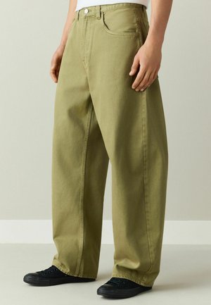 Person trägt locker sitzende olivgrüne Hose und schwarze Turnschuhe und steht vor einem schlichten hellen Hintergrund.