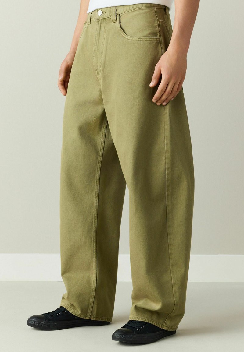 Personne portant un pantalon ample vert olive et des baskets noires, debout devant un fond clair uni.