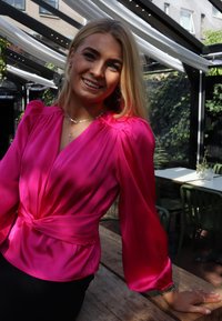 Svetlo roza svilena bluza z dolgimi napihnjenimi rokavi, globokim V-izrezom in pasom, ki se zaveže. Gladek tekstura in subtilen sijaj poudarjata dizajn.