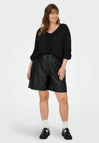 Smaidīga sieviete valkā melnu blūzi ar volāniem, melnas ādas šortus, baltas zeķes un melnus apavus, stāvot pret vienkāršu gaišu fonu.