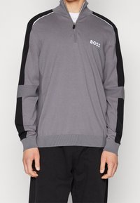 Mann trägt einen grauen Sweatshirt mit Viertelreißverschluss und schwarzen Ärmelstreifen sowie dem "BOSS"-Logo auf der Brust, kombiniert mit schwarzen Hosen.