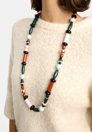 Długi naszyjnik z koralików w kolorach zielonym, czarnym, różowym, rdzawym, jasnoniebieskim i złotym, noszony na kremowym, teksturowanym swetrze.