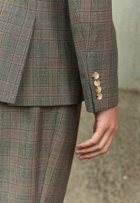 Šedý kostkovaný sako s oranžovými akcenty. Obsahuje čtyři béžové knoflíky na manžetách a texturovaný tkaný design. Přizpůsobený střih.