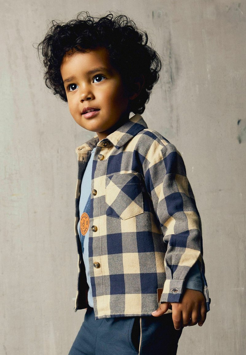 Jeune enfant aux cheveux bouclés portant une chemise à carreaux bleue et beige par-dessus un haut bleu, debout devant un fond gris clair uni.