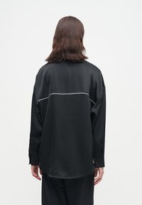 Άτομο με καστανά μαλλιά μέχρι τους ώμους φοράει loose μαύρο πουκάμισο με λευκή ρέουσα διακόσμηση στο πάνω μέρος της πλάτης, στέκεται μπροστά σε λευκό φόντο.