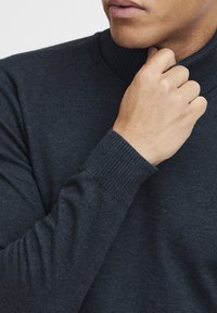Temno modra pulover s turtleneck iz mehke, teksturirane tkanine. Ima rebraste manšete in ovratnik za boljše prileganje. Povečan posnetek roke, ki prilagaja ovratnik.
