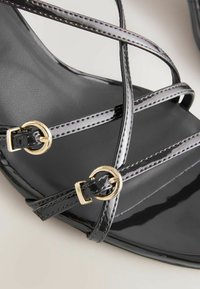 Sorte patentlæder sandaler med korslagte stropper, guld cirkelspænder og en glat tekstur. Har et elegant, moderne design.