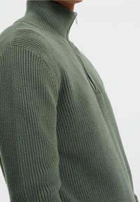 Maglione zippato a maglia in verde oliva. Presenta una texture a costine, colletto alto e zip laterale. Tessuto morbido e liscio con maniche aderenti.