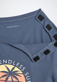 Μπλε ύφασμα με μαύρους ιμάντες βέλκρο στο γιακά, λευκή ετικέτα "Pier One" και μερική γραφική παράσταση φοίνικα με το κείμενο "Endless Sum".