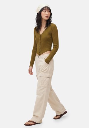Kvinde iført olivengrøn cropped ribstrikket cardigan, beige cargobukser, cremefarvet tørklæde og brune sandaler, som går med den ene hånd i lommen.