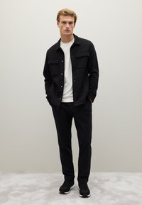 Giacca camicia nera con due tasche sul petto, indossata su una t-shirt bianca. Abbinata a pantaloni neri e scarpe da ginnastica nere.
