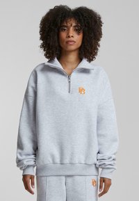 Lysegrå hettegenser med halv-glidelås krage, ribbestrikkede mansjetter og en oransje brodert logo på brystet. Mykt, teksturert stoff.