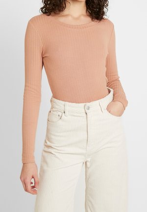 Femme portant un haut pêche côtelé à manches longues rentré dans un pantalon blanc côtelé taille haute, une main dans la poche.