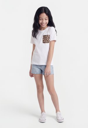 Fată zâmbitoare cu păr lung și întunecat poartă un tricou alb cu buzunar cu imprimeu de leopard, pantaloni scurți de denim deschis și adidași albi, stând pe un fundal alb.