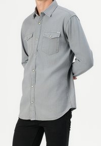 Camisa de mezclilla gris con mangas largas, cierre frontal de botones, dos bolsillos en el pecho y detalles de costura sutiles. Combinada con pantalones negros.