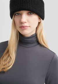 Grauer Rollkragenpullover aus glattem Stoff mit einer körpernahen Passform. Schwarze gerippte Strickmütze auf hellblonden Haaren. Natürlicher Hautton.