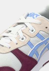 Γκρι αθλητικά παπούτσια με λευκές κορδόνες, μπεζ και μπλε λεπτομέρειες, και βουργουνδί σουέτ λεπτομέρεια στο πλάι. Διαθέτει υφή στην επάνω πλευρά και καουτσούκ σόλα.