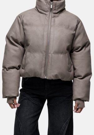 Veste matelassée taupe avec col montant et fermeture éclair devant, portée avec un pantalon noir, montrant des manches longues avec des trous pour le pouce.