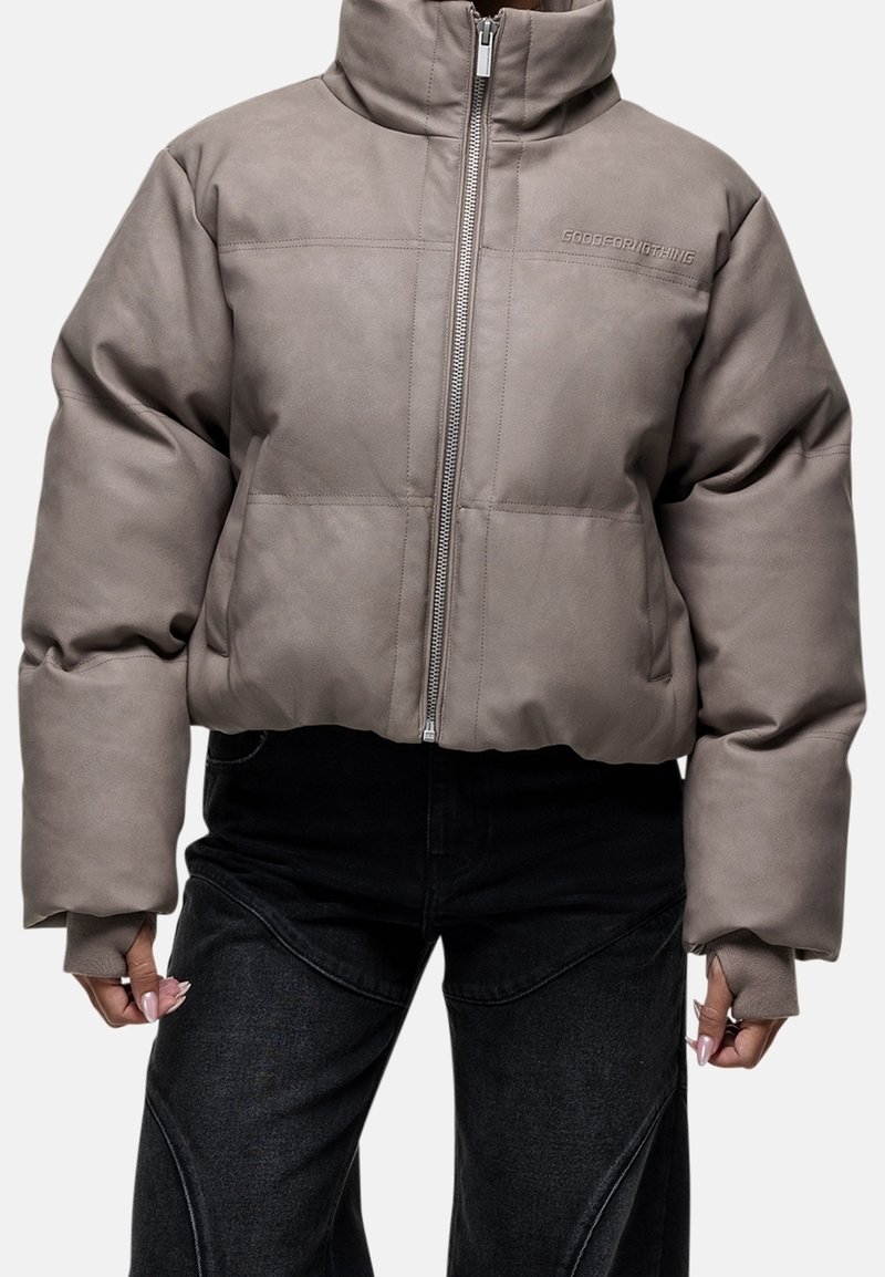 Veste matelassée taupe avec col montant et fermeture éclair devant, portée avec un pantalon noir, montrant des manches longues avec des trous pour le pouce.
