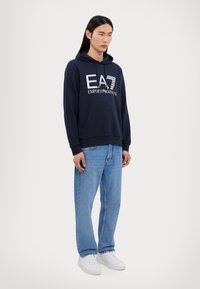 Felpa in cotone blu navy con tasca frontale, logo bianco "EA7 EMPORIO ARMANI," abbinata a jeans in denim blu chiaro e scarpe bianche.