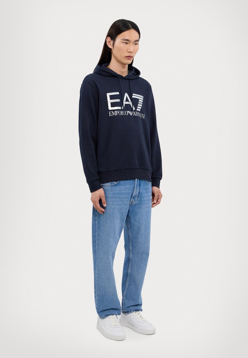 Felpa in cotone blu navy con tasca frontale, logo bianco "EA7 EMPORIO ARMANI," abbinata a jeans in denim blu chiaro e scarpe bianche.