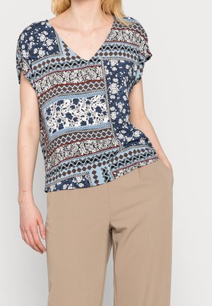 Mujer que lleva una blusa de manga corta con cuello en V y patrones mixtos florales y geométricos en azul, blanco y marrón, combinada con pantalones beige.
