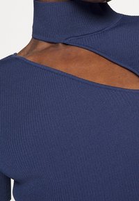 Marinblå ribbad stickad topp med en hög krage, asymmetrisk utskärning och mjuk textur med synliga sömmar.