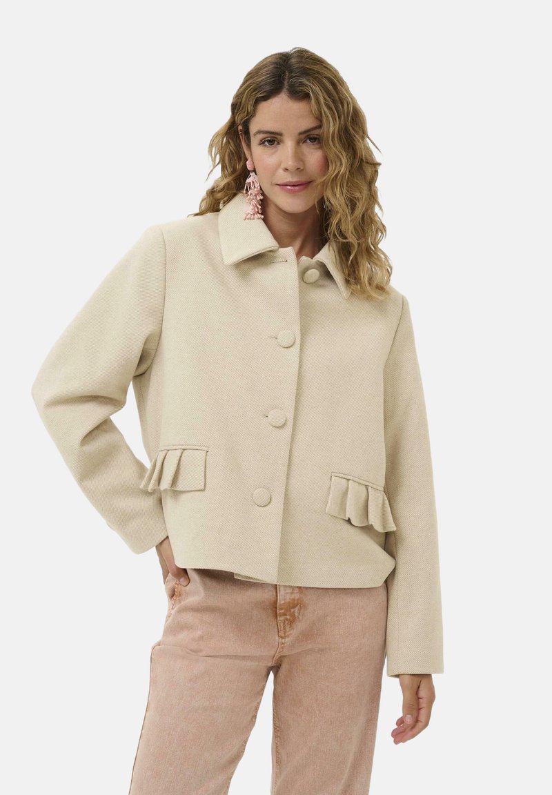 Beige cropped dzseki strukturált anyagból, gallérral, elülső gombokkal és fodros zsebekkel. Világos rózsaszín nadrággal viselve.