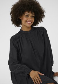 Chemise noire à boutons avec un col haut, des coutures d'épaules froncées et des poignets élastiques. Tissu doux avec une coupe loose, design minimaliste.