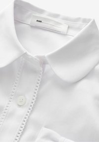 Camicia bianca con colletto, patta dei bottoni rifinita in pizzo e etichetta cucita con la scritta "NOME" e linee vuote per scrivere.