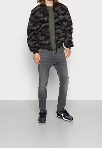 Chaqueta bomber de camuflaje negro y gris, camiseta verde, vaqueros grises, zapatillas negras con detalles blancos, mostrada de pie sobre un fondo neutro.
