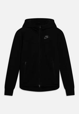 Melnā Nike kapuces jakā ar kapuci, ar rāvējslēdzēja priekšpusi un slēptām kabatām. Izgatavota no mīksta, teksturēta auduma.